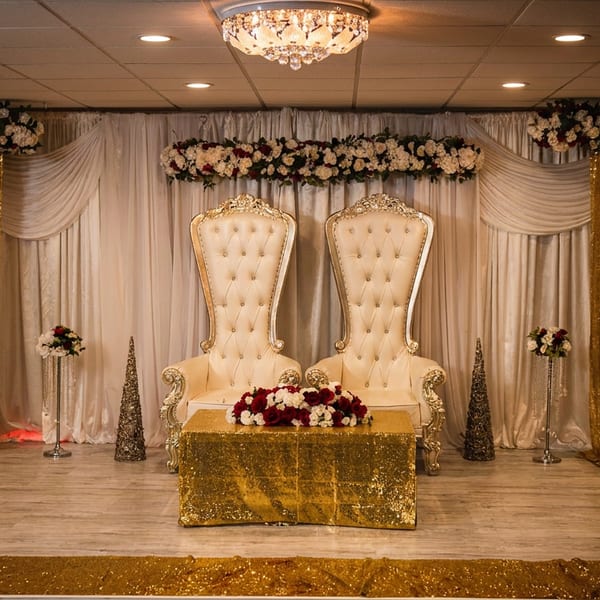 Elegant stage with two ornate white chairs, a gold sequin table, and floral arrangements.