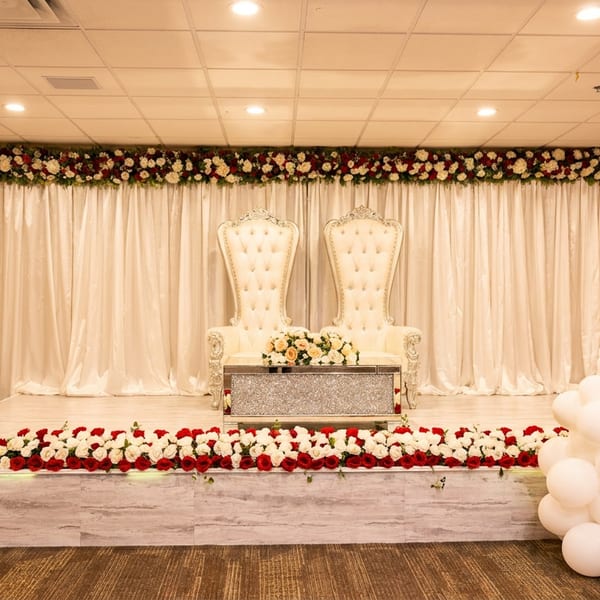 Elegant stage setup with two ornate white chairs, floral arrangements, and red and white balloons.