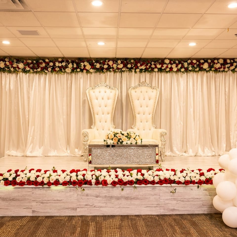 Elegant stage setup with two ornate white chairs, floral arrangements, and red and white balloons.