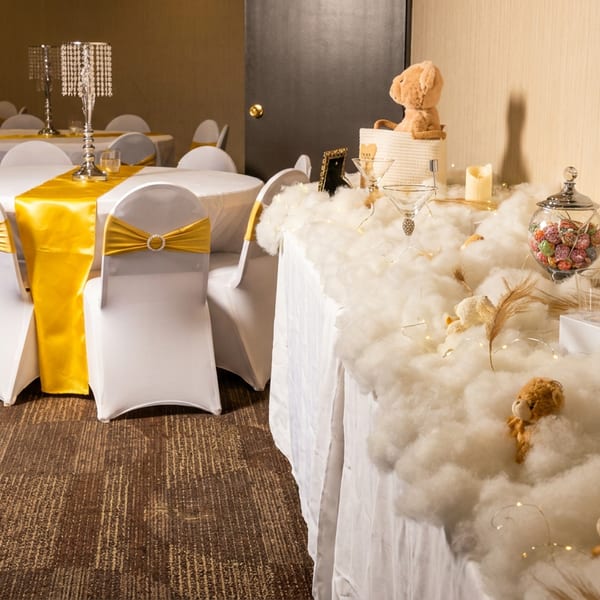 A banquet hall with round tables draped in white and yellow, and a dessert table with cotton-like decor.