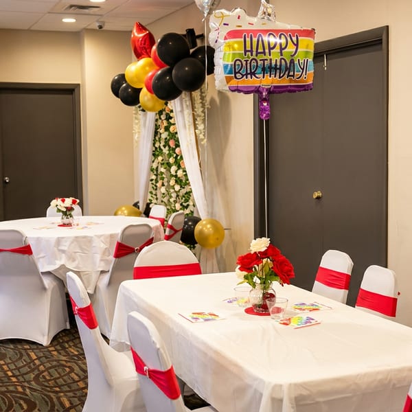 A decorated room with white tables and chairs with red sashes, balloons, and a "Happy Birthday" sign.