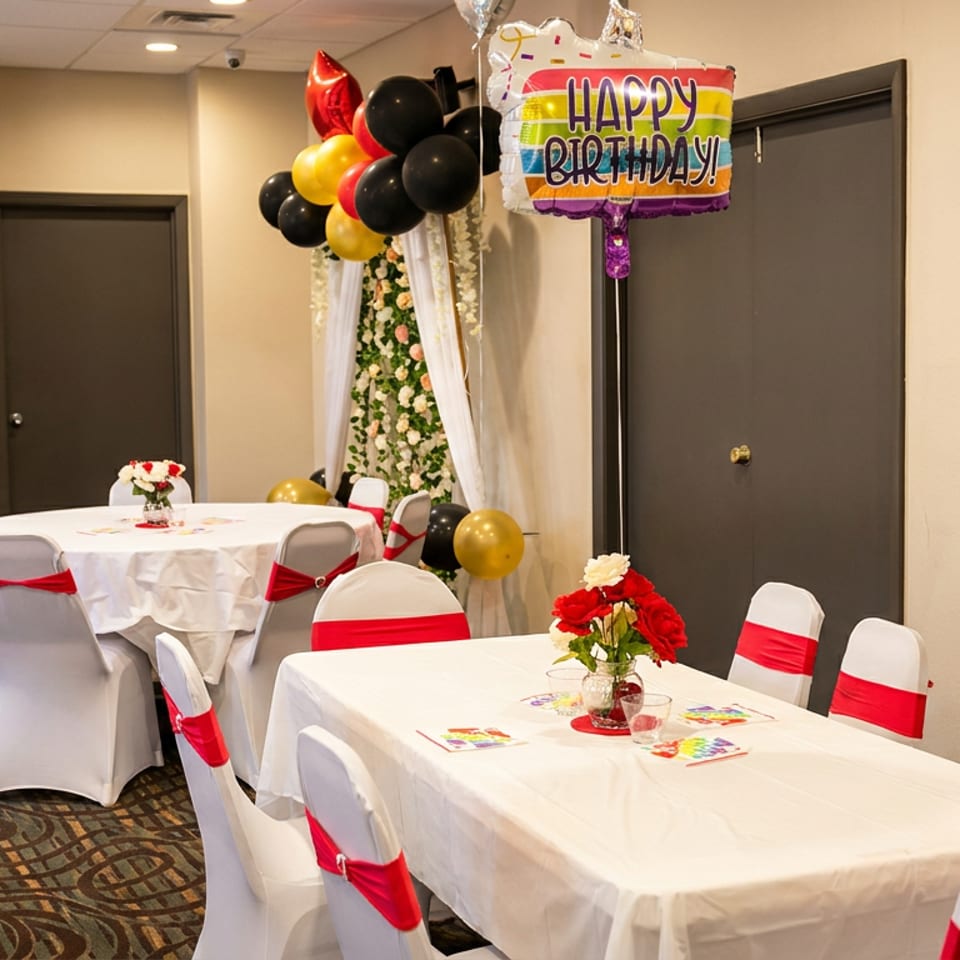 A decorated room with white tables and chairs with red sashes, balloons, and a "Happy Birthday" sign.