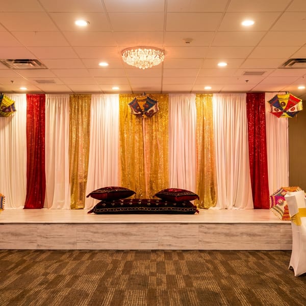 A decorated stage with red, white, and gold curtains, floor cushions, and colorful umbrellas in a banquet hall.