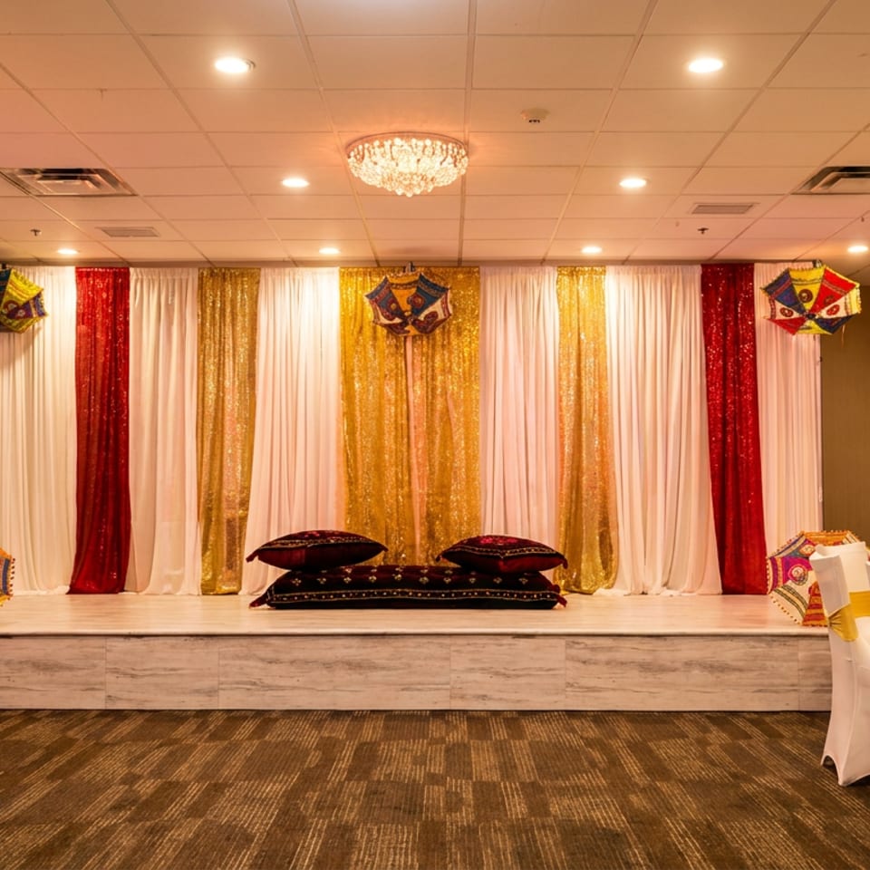 A decorated stage with red, white, and gold curtains, floor cushions, and colorful umbrellas in a banquet hall.